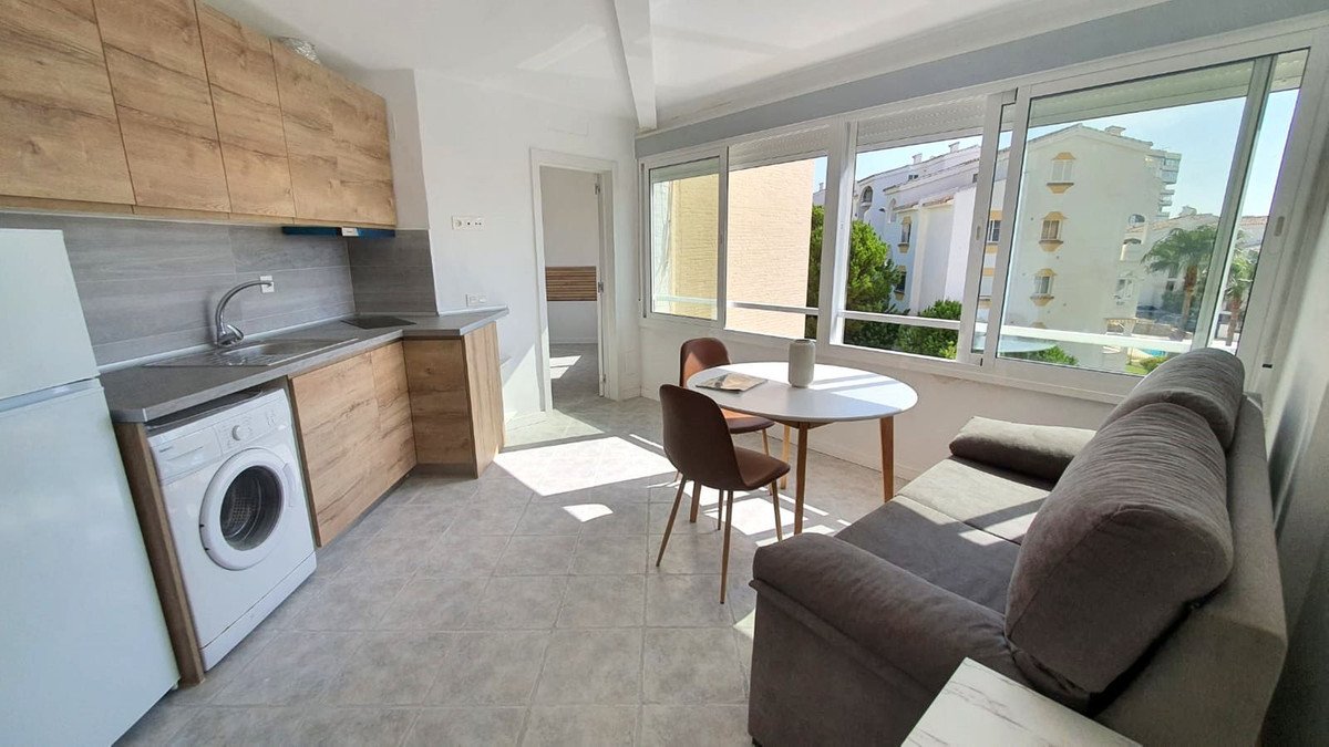 Modern kitchen-living area with wooden cabinets, washing machine, and large windows overlooking buildings and greenery.