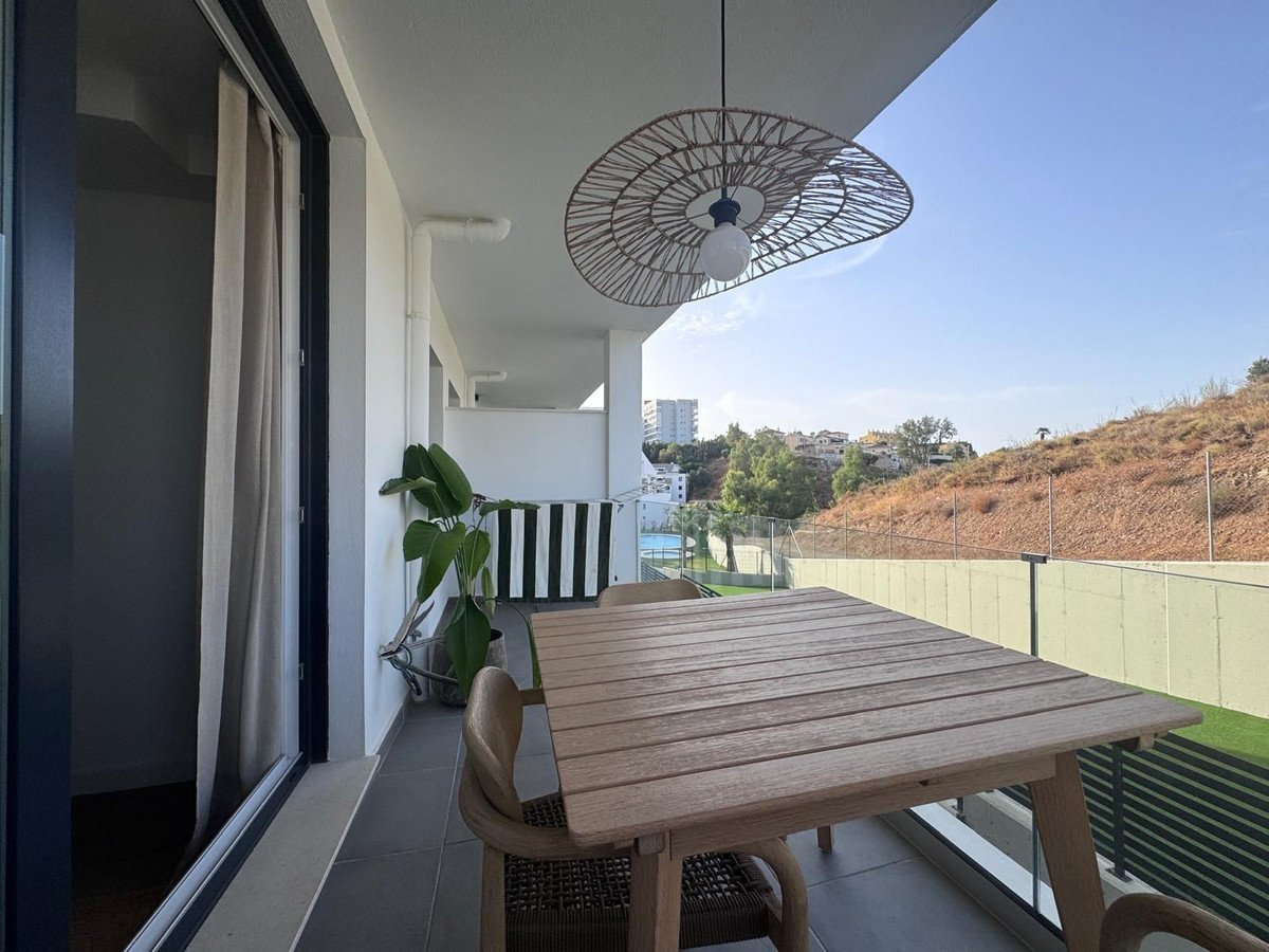 A wooden dining table and chairs on a balcony with a view of a hillside, buildings, and greenery.