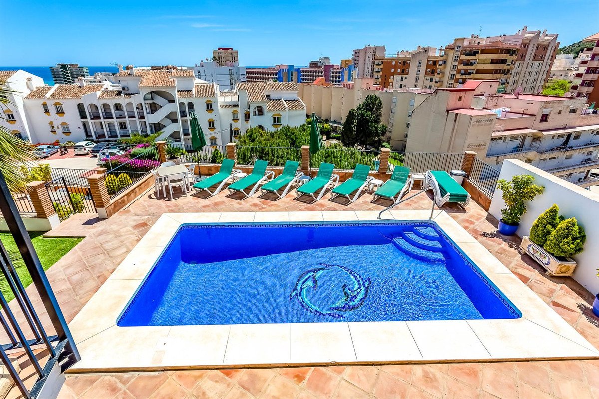 Rooftop terrace with a swimming pool, sun loungers, and a distant view of the sea and buildings.