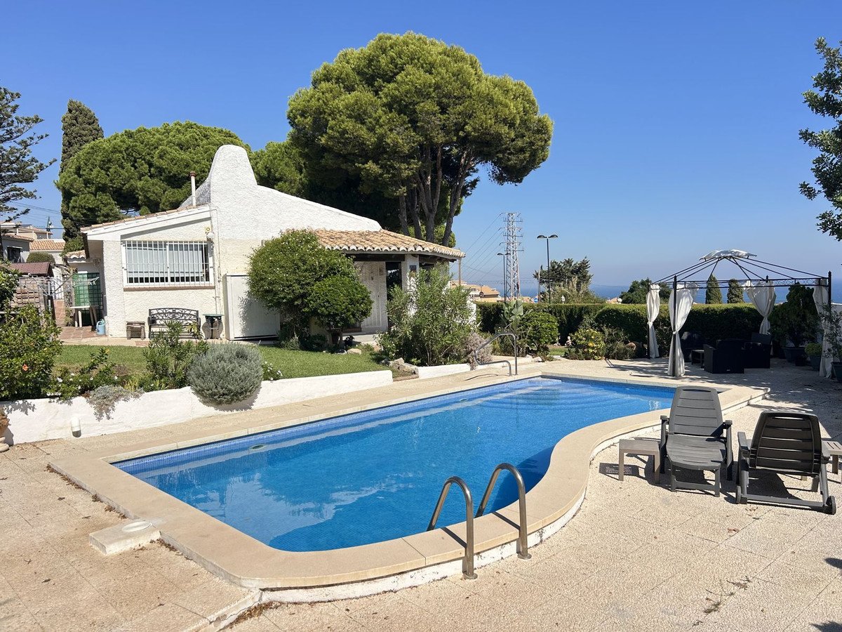 A villa with a swimming pool, lounge chairs, and a gazebo with a distant view of the sea on a sunny day.