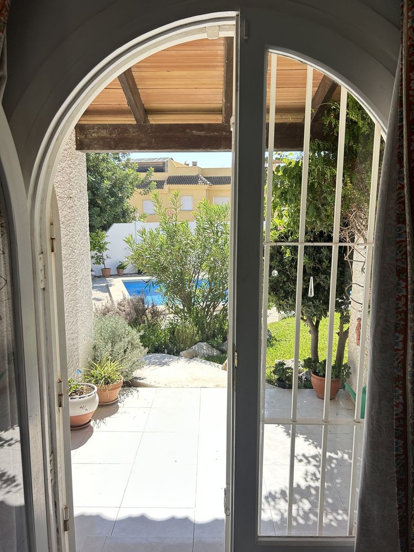 View from doorway to a tiled patio, garden with plants, swimming pool, and houses beyond.