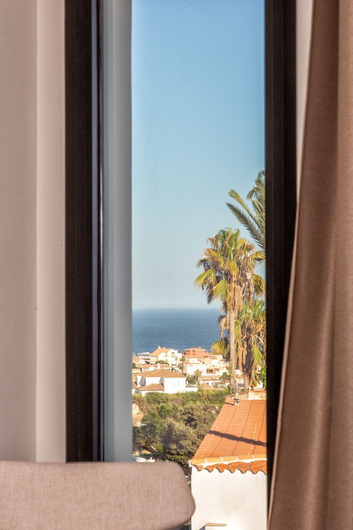 View of the Mediterranean Sea, coastal villas, and palm trees seen through a window with curtains.