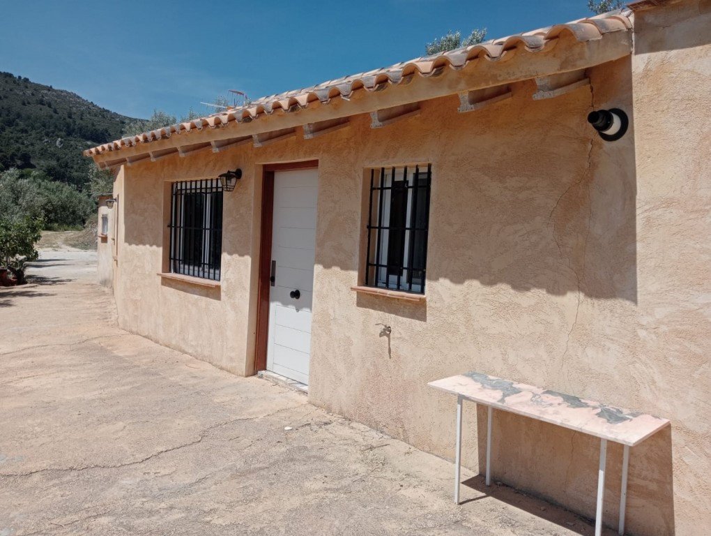 Exterior of a small cottage with barred windows, a white door, and a console table on the patio, with hills in the background.