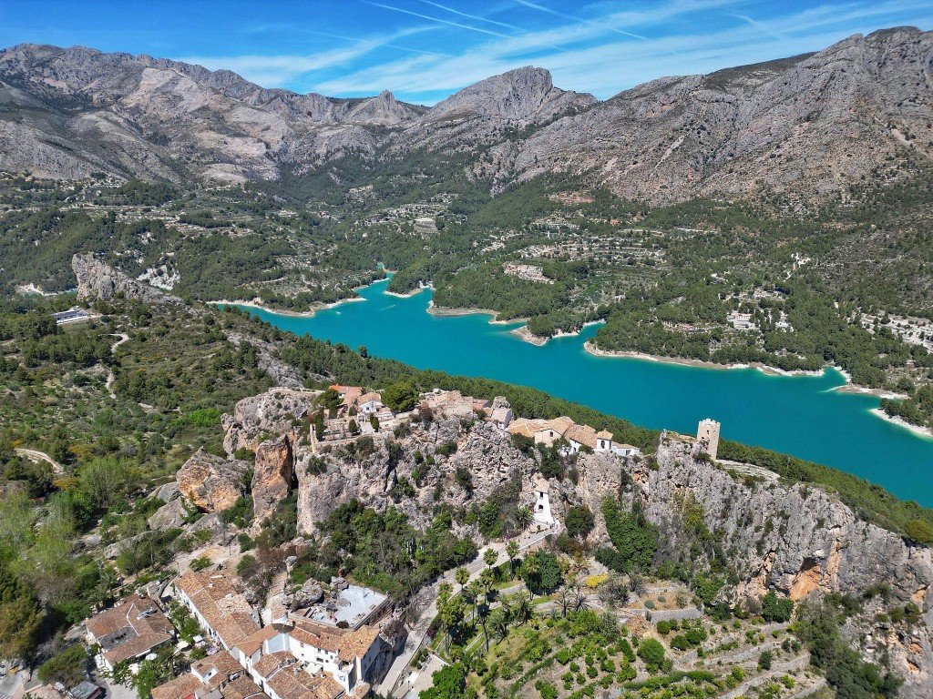 Aerial view of a historic village nestled on a rocky cliff overlooking a turquoise lake and surrounding mountains.