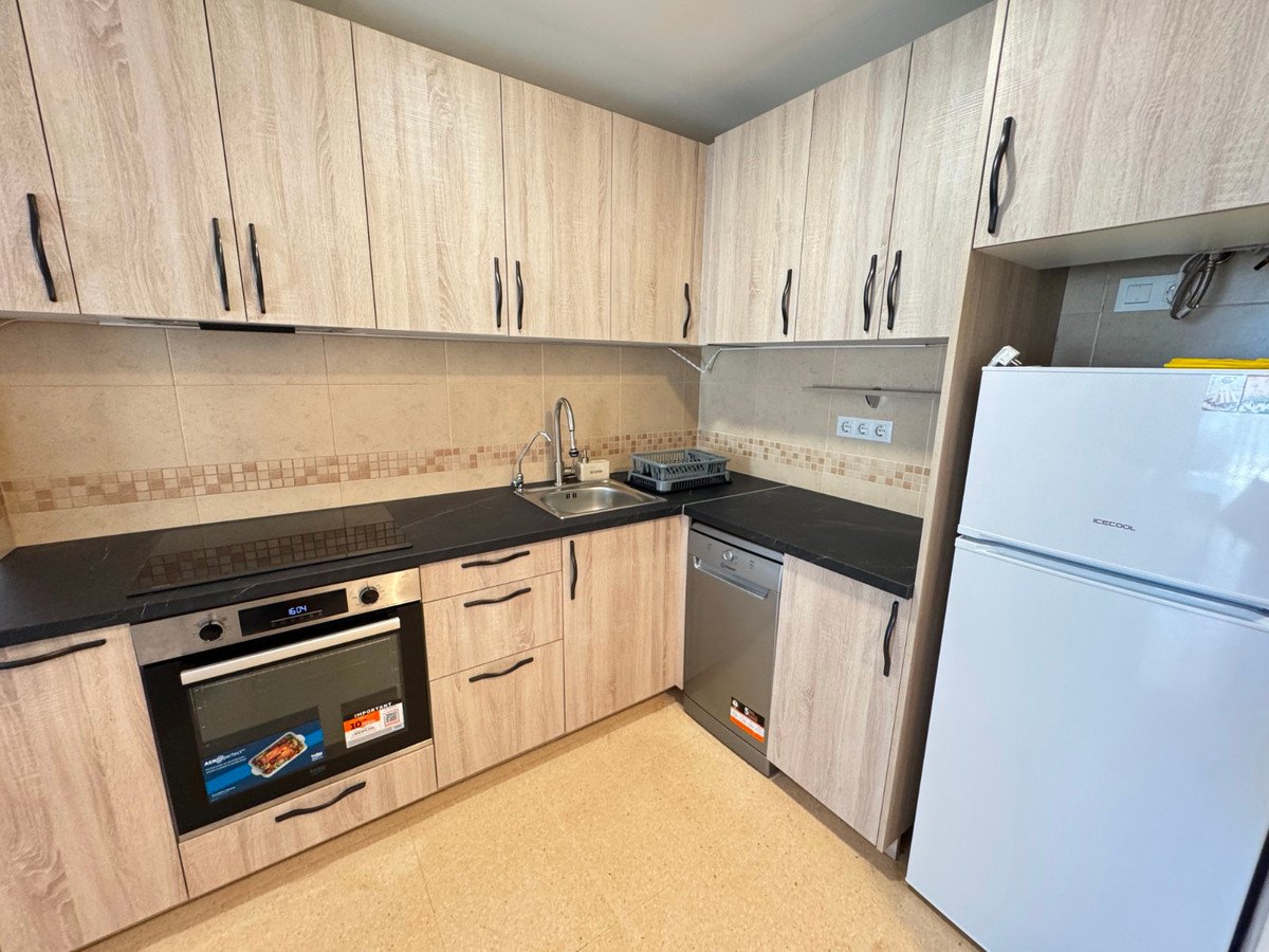 A modern kitchen with light wood cabinets, black countertops, oven, dishwasher, sink, and a white refrigerator.