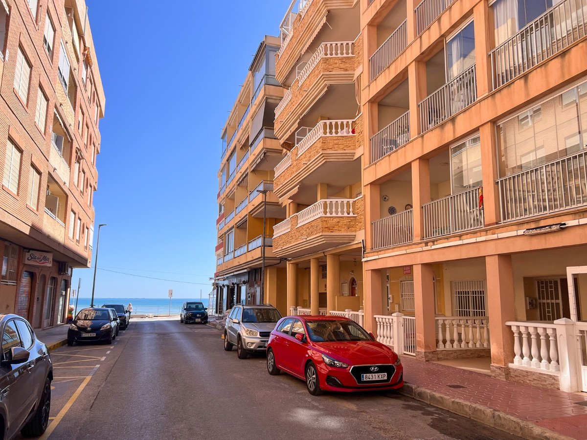 Street view of apartment buildings with balconies. The sea is visible at the end of the street under a clear blue sky.