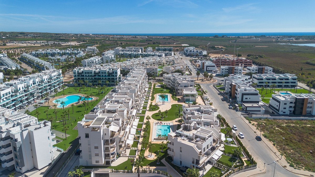 Aerial view of modern white apartment buildings with swimming pools and manicured lawns, overlooking the sea in the distance.