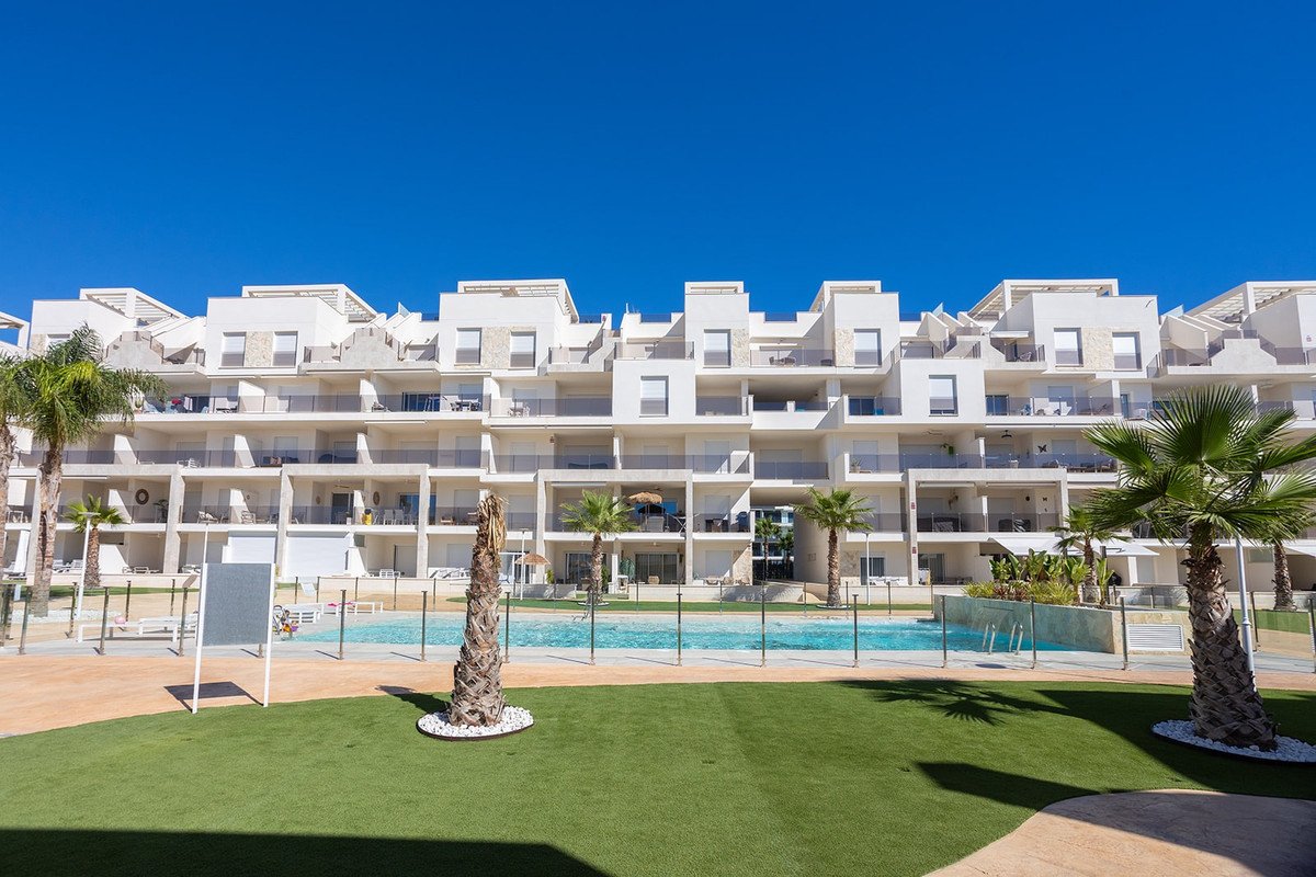 Modern white apartment building with balconies overlooking a swimming pool and artificial grass courtyard with palm trees.
