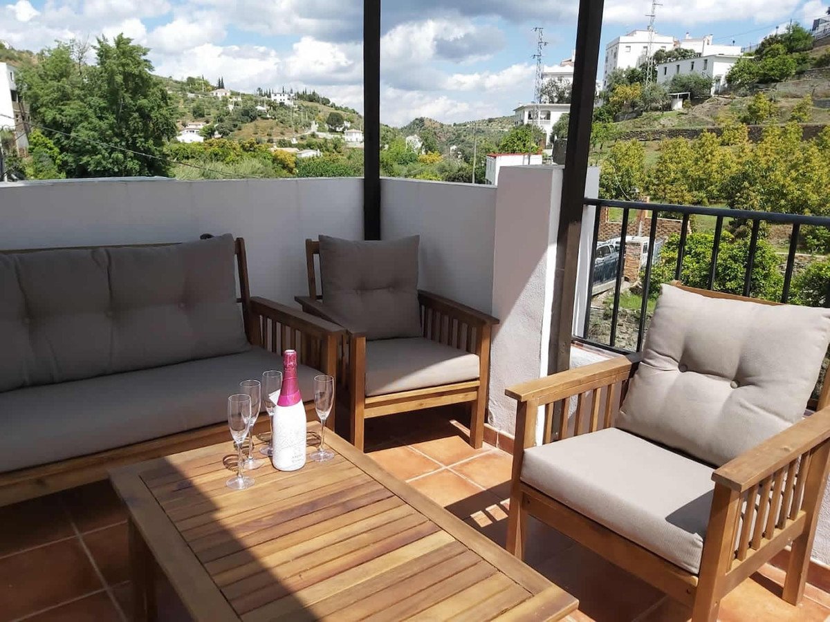 Wooden terrace furniture with champagne glasses and bottle, overlooking rolling hills and white houses.
