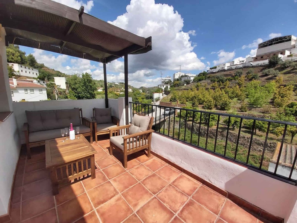 A covered terrace with wooden seating and a coffee table overlooks a sunny hillside with trees and white houses.