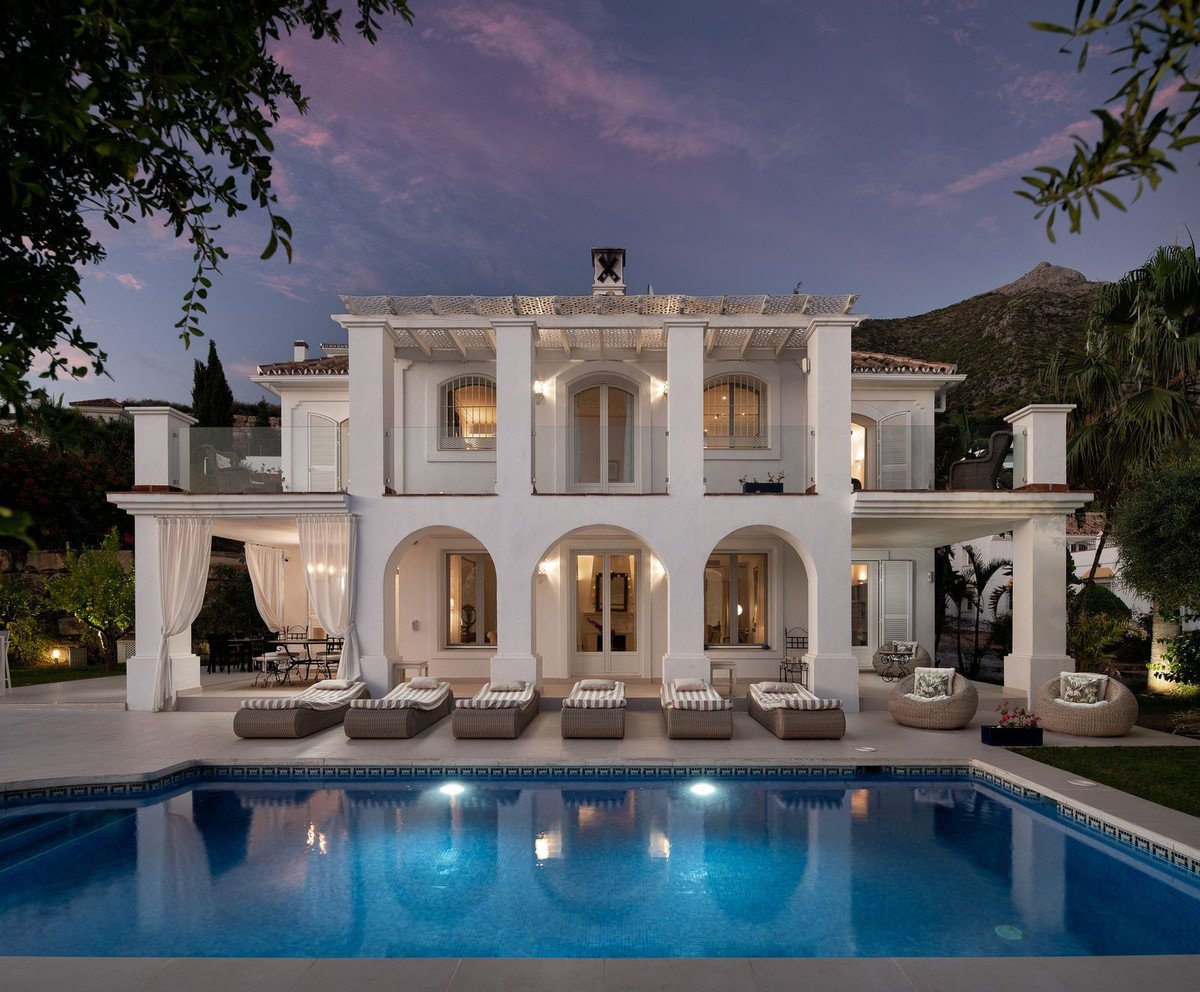 Luxury white villa with arched windows, a swimming pool, lounge chairs, and a scenic mountain view at sunset.