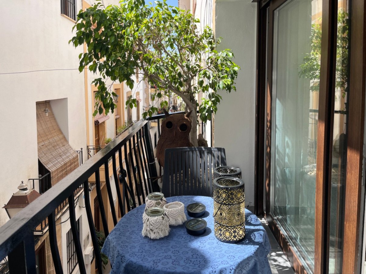 A small balcony with a table, chairs, a plant, and views of a narrow street with buildings.