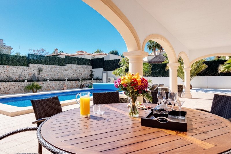 Outdoor dining table and chairs set up under a covered patio with a view of a swimming pool and lush greenery.