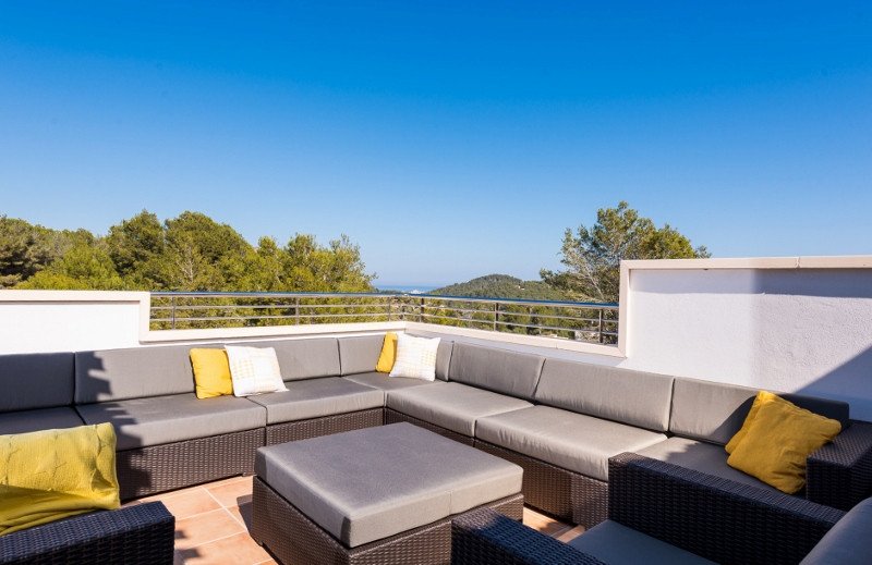 Spacious outdoor terrace with a large sectional sofa, coffee table, and scenic views of trees and the sea under a clear blue sky.