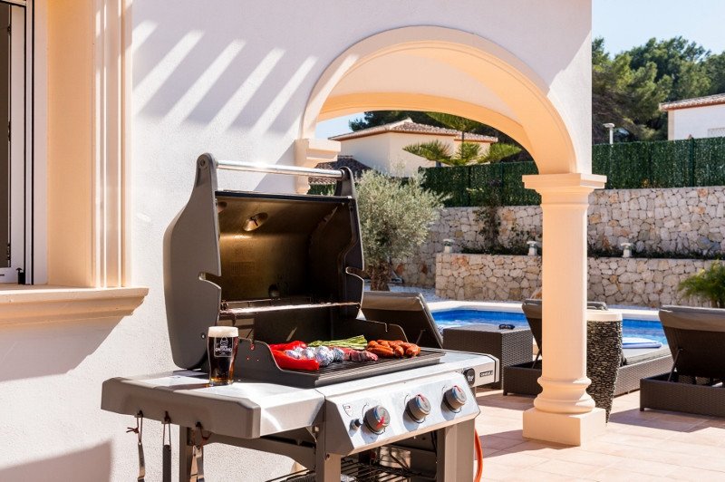 A stainless steel outdoor grill with food cooking on it, placed on a patio near a swimming pool and lounge chairs.