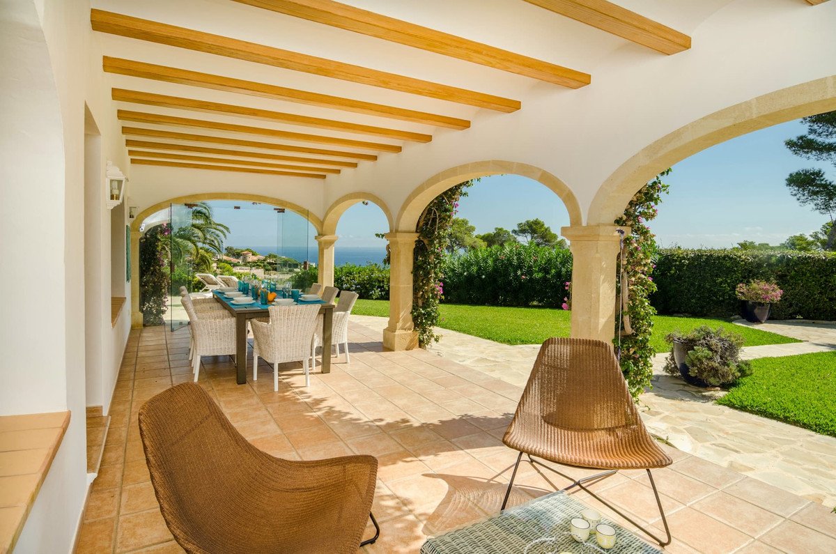 Covered terrace with dining table and chairs overlooking a lush green garden and the sea. Furnished outdoor seating area.