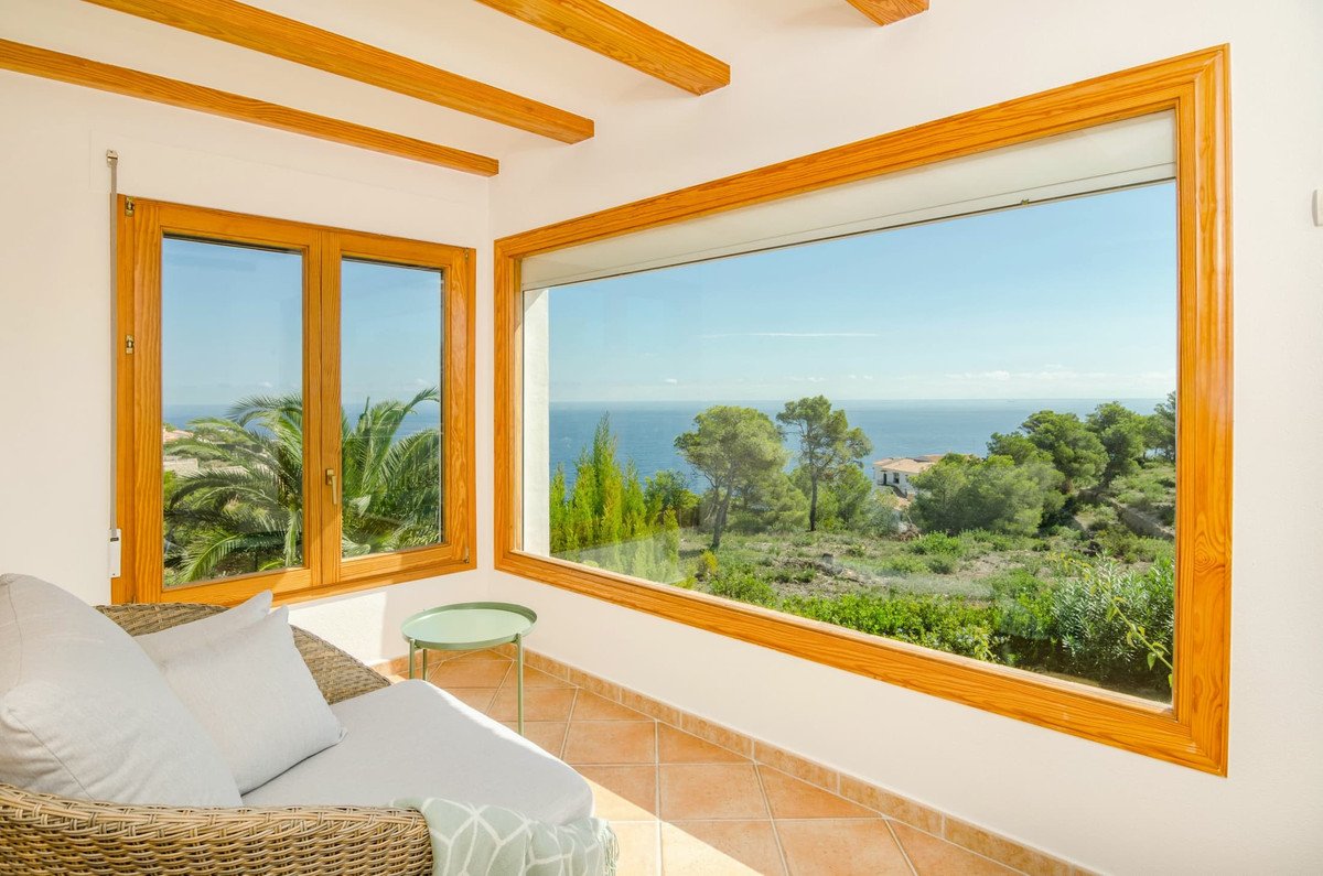 Sunroom with large windows offering stunning ocean views, palm trees, and lush greenery. Includes a lounge chair and side table.