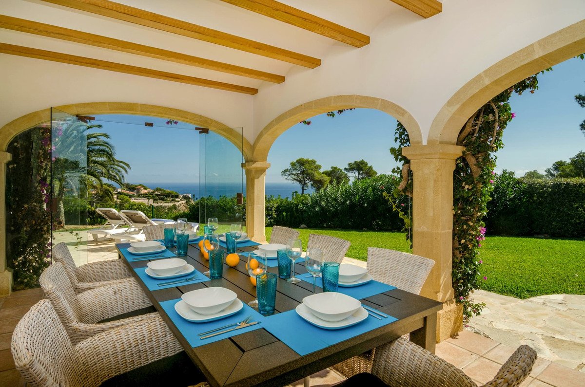A covered terrace with a dining table set for a meal, overlooking the ocean and lush green gardens.