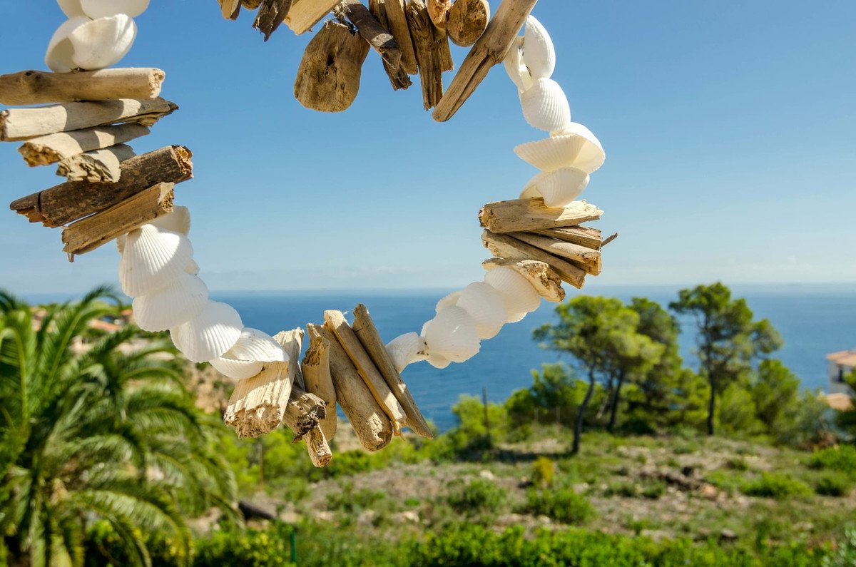 A handmade garland of shells and driftwood hangs against a backdrop of the blue ocean and lush green trees.
