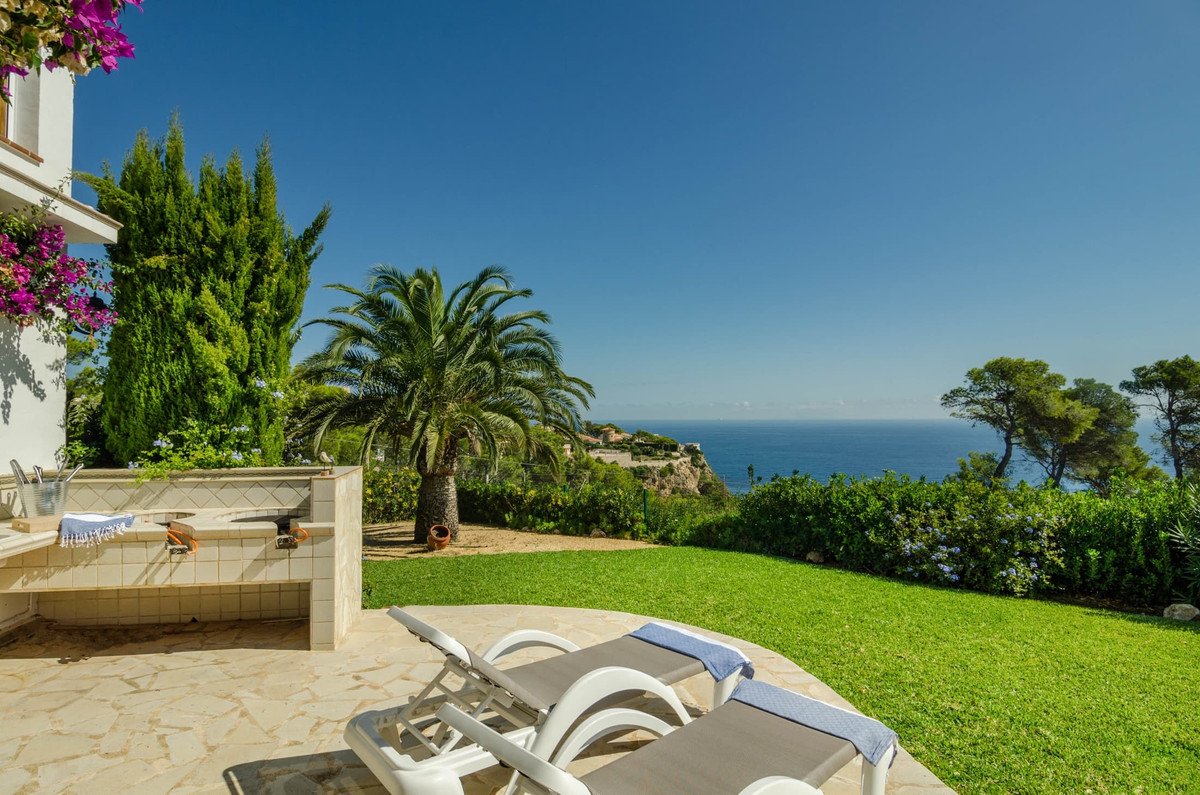 Outdoor patio with lounge chairs, built-in BBQ, green lawn, palm trees, and a stunning sea view.