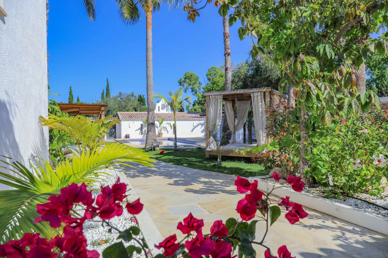 Overzicht van een weelderige tuin met een cabana en een wit gebouw op de achtergrond.