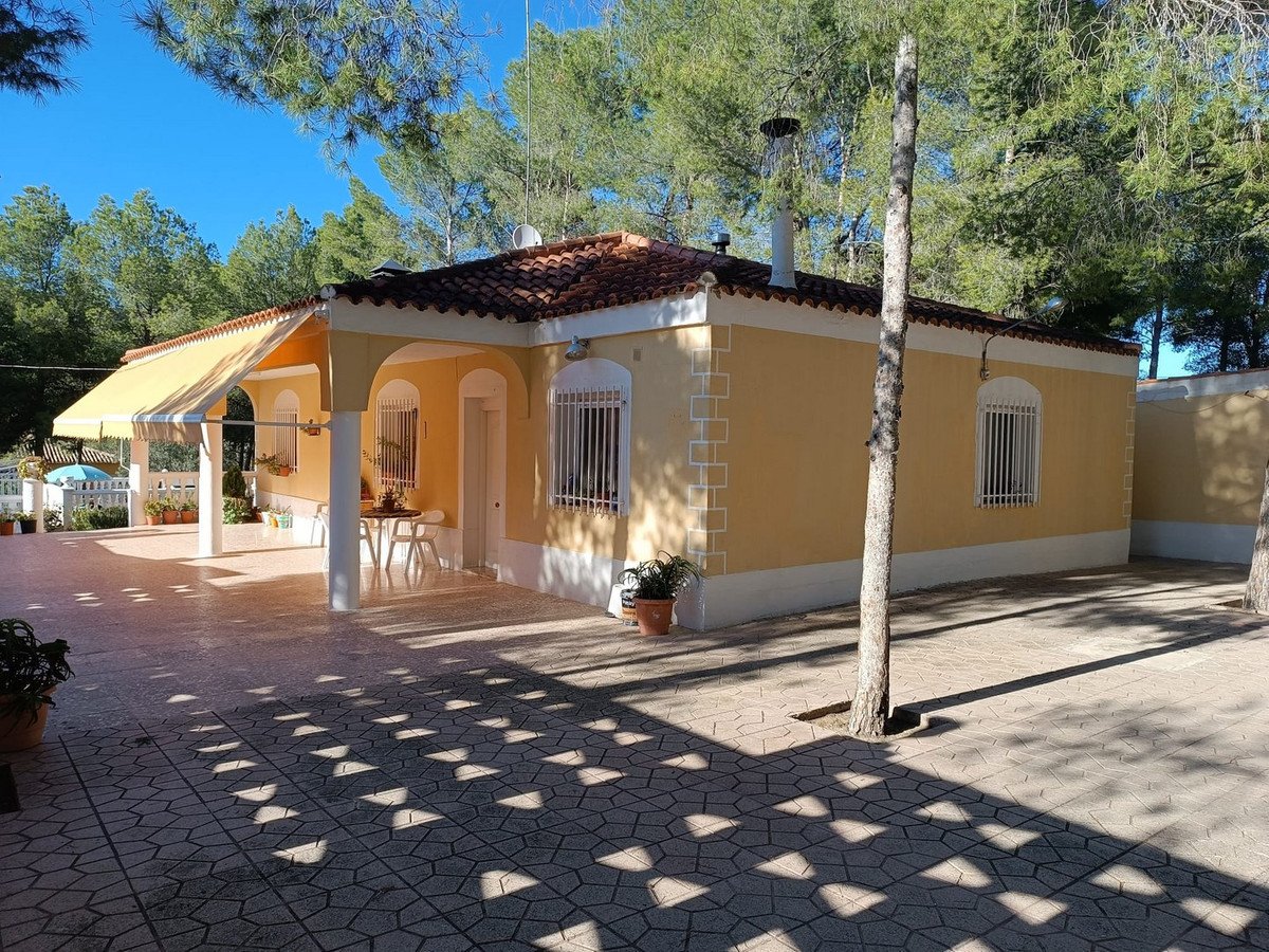 Yellow villa with white awning, outdoor seating area, and paved patio surrounded by pine trees.
