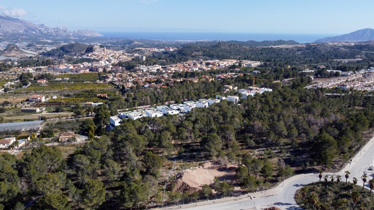 Aerial view of a modern townhouse development nestled in a forest, with a distant town and sea visible.