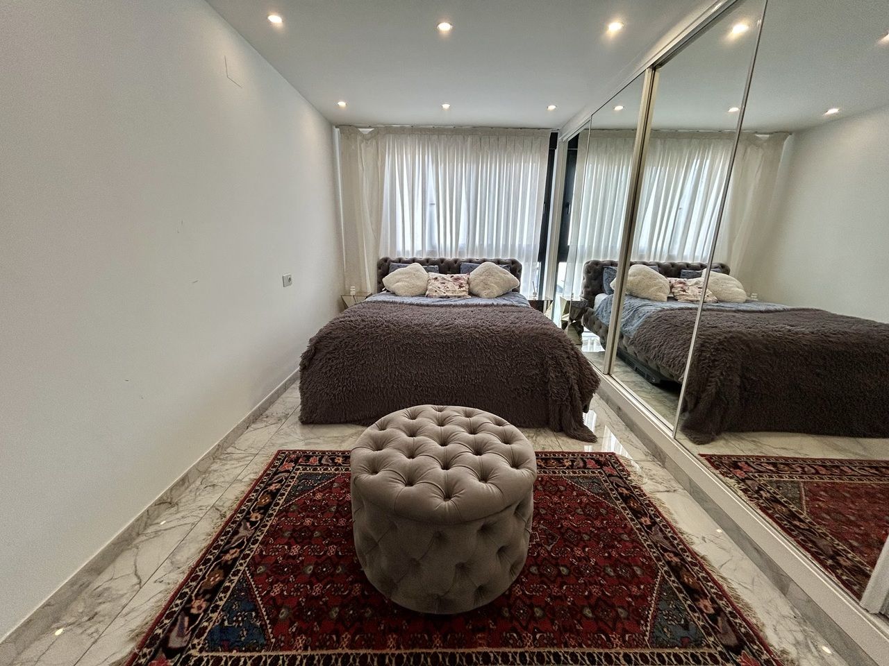 Stijlvolle slaapkamer met grijze poef, spiegelwand en bed.