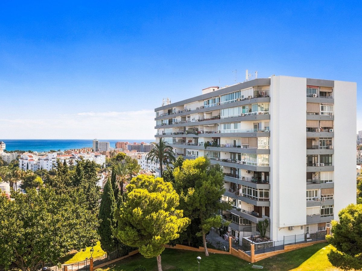 Modern apartment building with balconies overlooking the sea and town, surrounded by green trees under a clear blue sky.