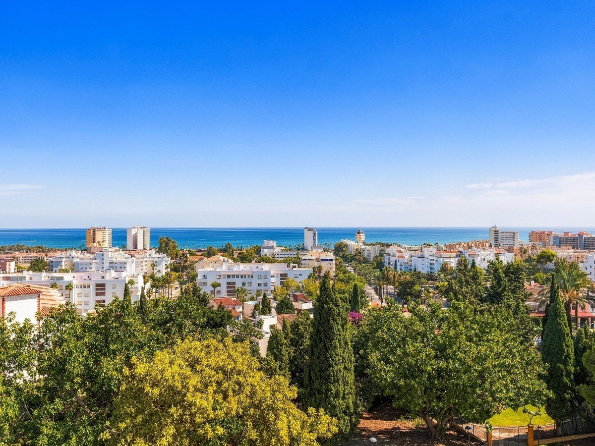 View from a balcony overlooking a town with trees and the sea in the distance under a clear blue sky.