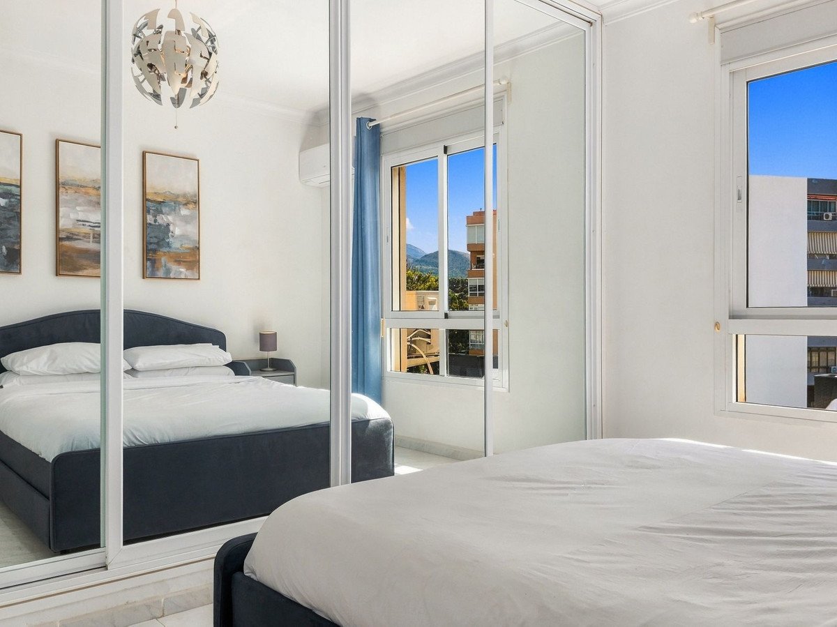 Bedroom with a large bed, mirrored closet doors reflecting the room and paintings, and a window with a mountain and cityscape view.
