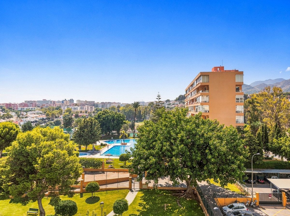 Apartment building with balconies overlooking a swimming pool, trees, and distant cityscape with mountains.