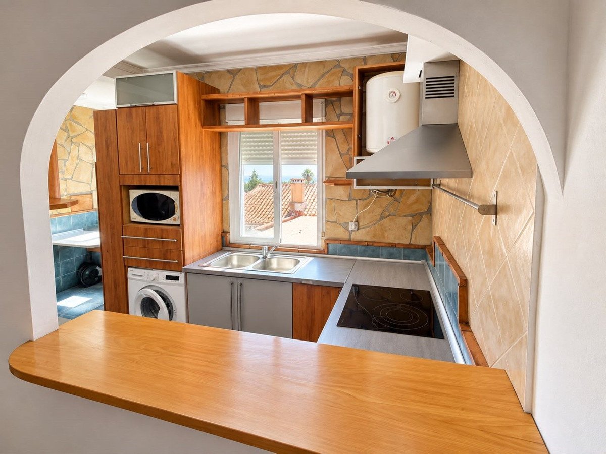 A modern kitchen with wooden cabinets, microwave, washing machine, electric hob, and a window overlooking rooftops and outdoor view.