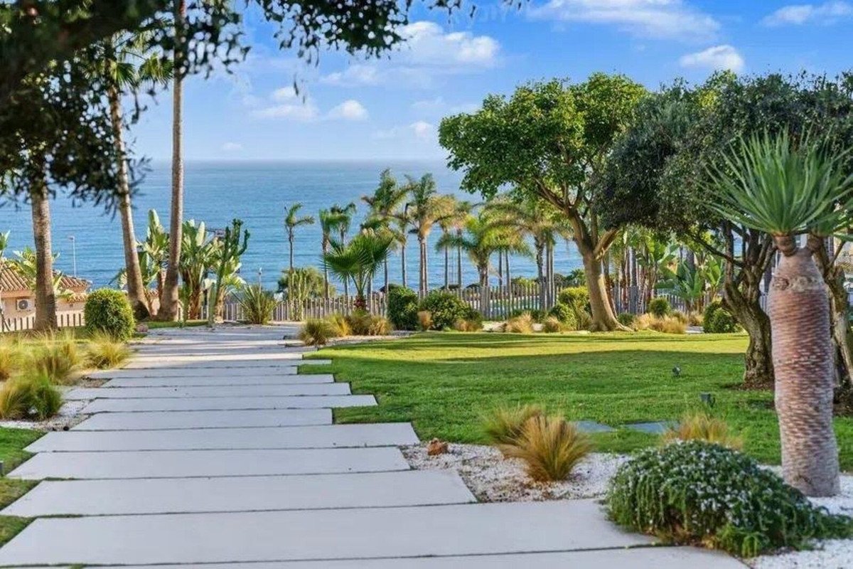 A modern pathway leads through a lush garden towards a beautiful sea view with palm trees and clear blue sky.