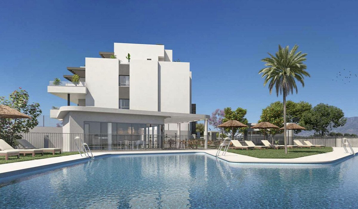 Modern apartment building with balconies, surrounded by a swimming pool, lounge chairs, umbrellas, and palm trees under a clear blue sky.