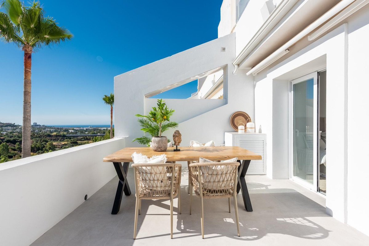 Terrace with a wooden dining table, chairs, and a view of the sea and palm trees under a clear blue sky.