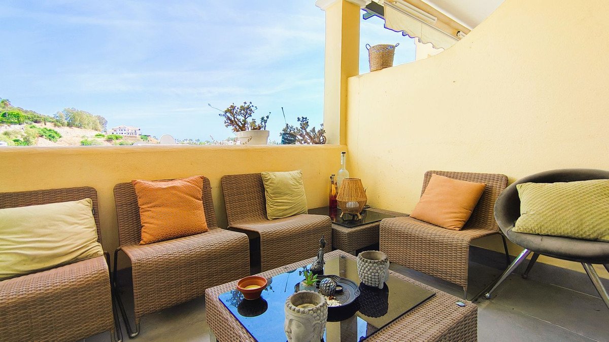 A sunny terrace with wicker chairs and a coffee table offering a view of a green hillside and buildings under a blue sky.