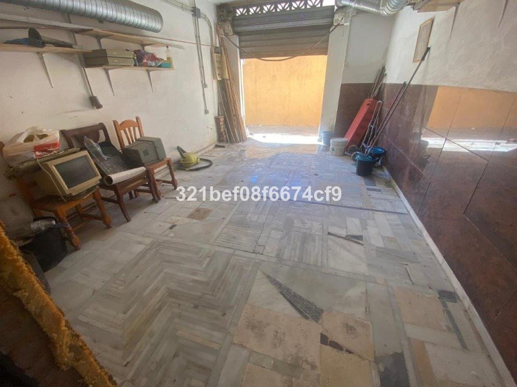 A cluttered storage area with an old computer, chairs, a vacuum cleaner, and tools near an open garage door.