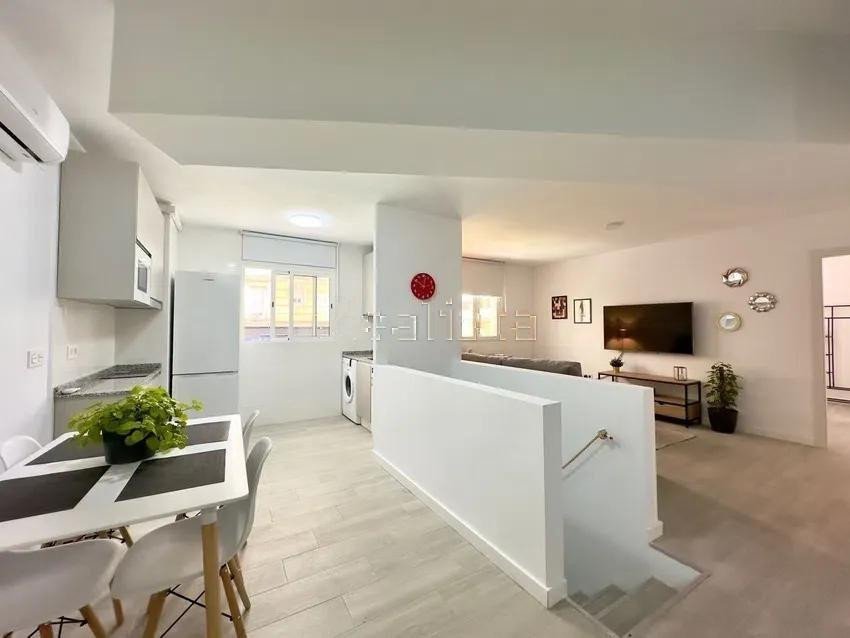 Modern apartment interior with open-plan living, dining, and kitchen area, featuring a dining table and sofa with TV.