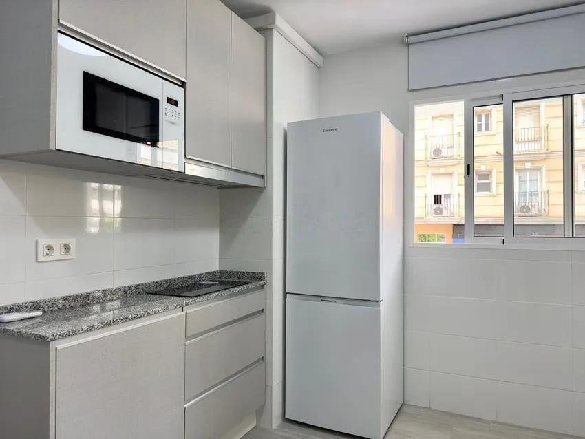 Modern white kitchen with granite countertops, stainless steel microwave, refrigerator, and view of building exterior through window.