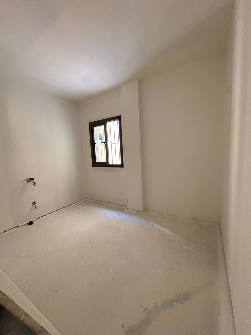 An empty room with a window and exposed plumbing fixtures. The walls are painted white and the floor is bare concrete.