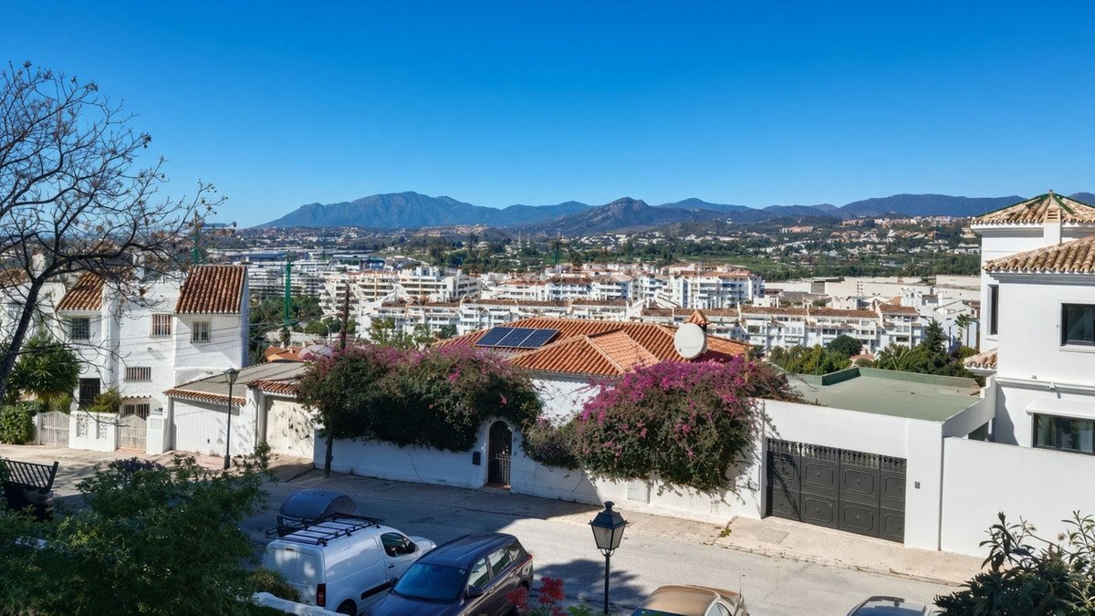 A view of a villa with purple flowers in front, overlooking a city and mountains under a clear blue sky.