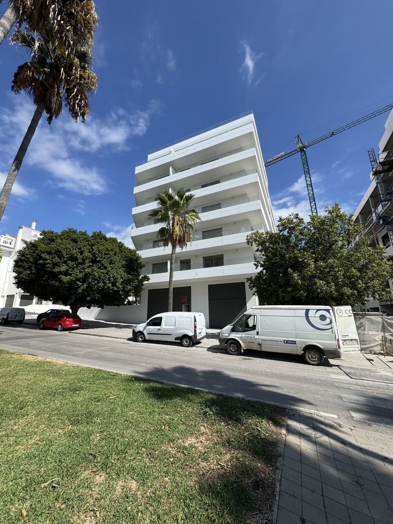 Modern white apartment building with balconies, palm trees, and construction crane under a blue sky.
