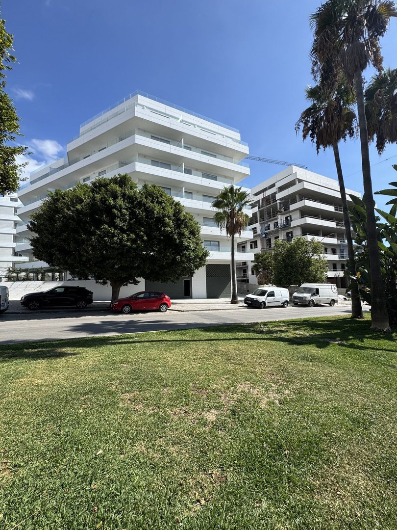 Modern white apartment building with multiple balconies, surrounded by greenery and palm trees on a sunny day.