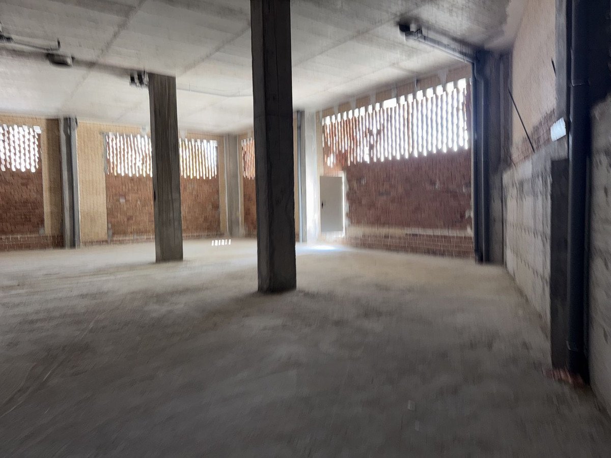 Unfinished industrial space with concrete floor, brick walls, and large pillars. Natural light enters through narrow windows.