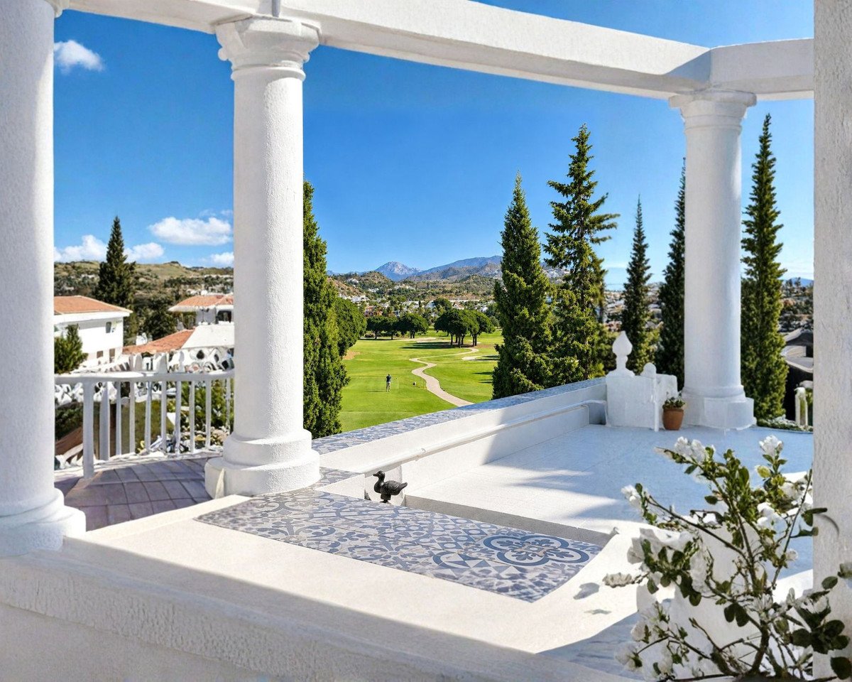 Balcony with columns overlooking a green golf course, resort buildings, and mountains under a blue sky.
