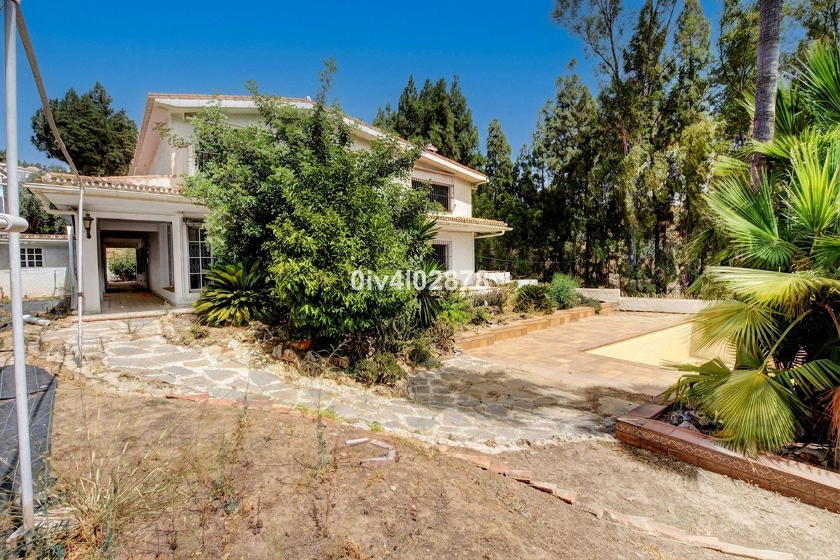 Exterior view of a white villa with a covered walkway, surrounded by lush greenery, trees, and a paved patio area.