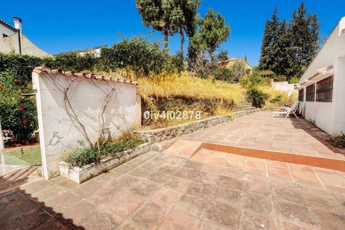 A tiled terrace with steps leads up to a garden area and the side of a white house under a clear blue sky.