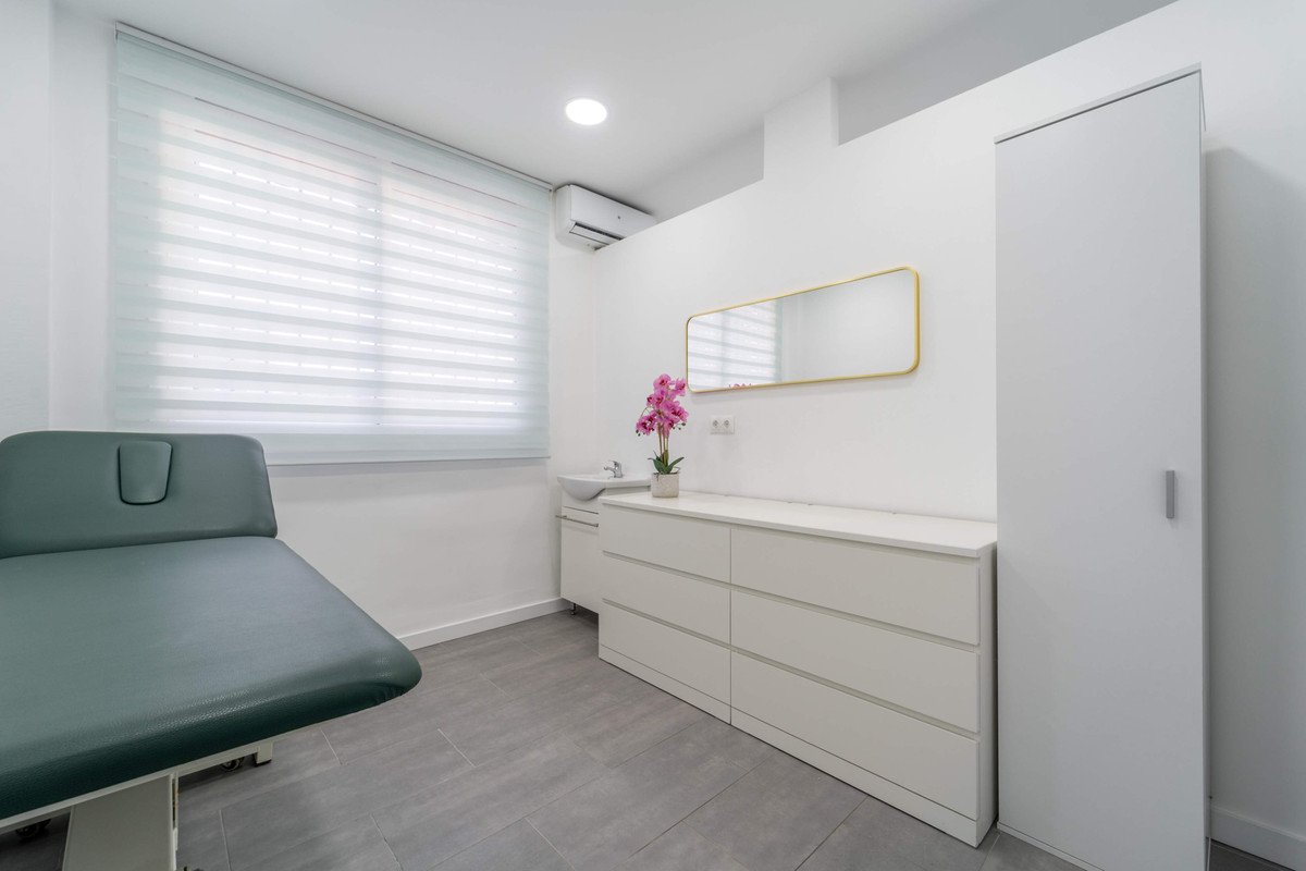 A modern medical examination room with a window, green bench, and white cabinets.