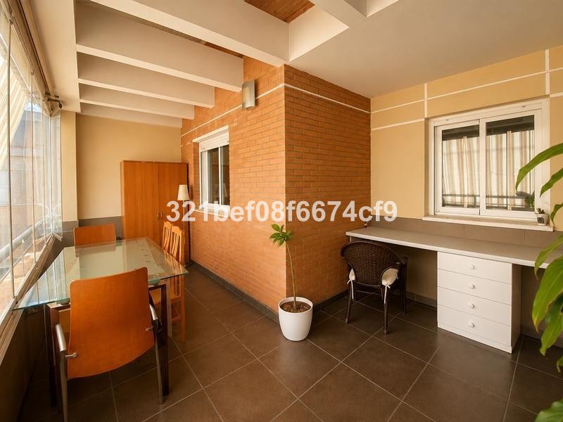 Balcony area with a glass dining table, chairs, a desk with drawers, and a wicker chair. There's a window with curtains.